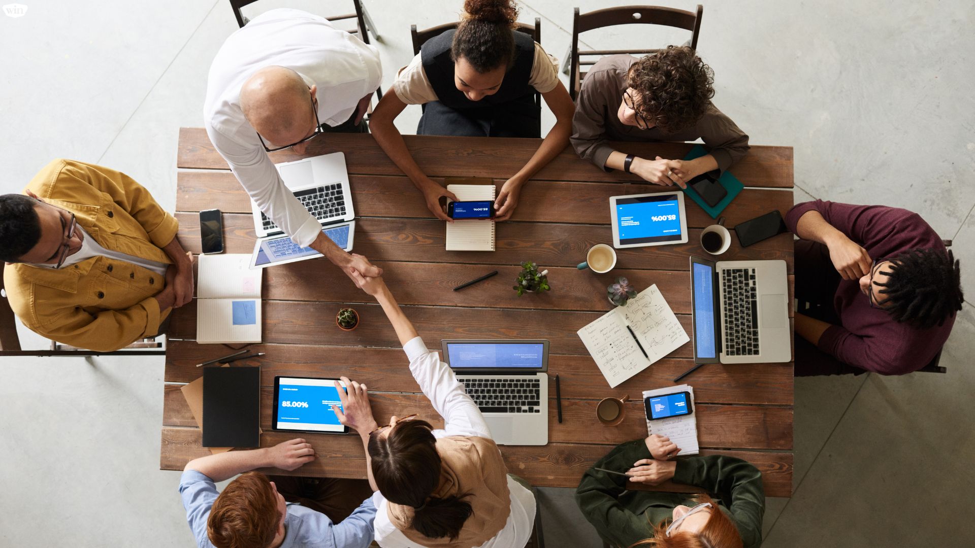 Team meeting around a table working on their endpoint devices. Two professionals are shaking hands across the table.