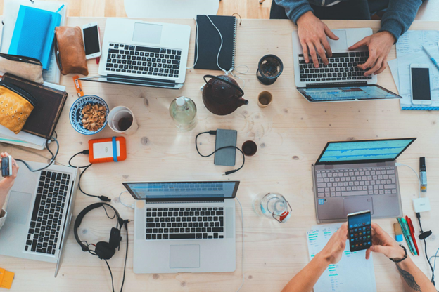 Several laptops and electronics are on a table, with people sitting around working.
